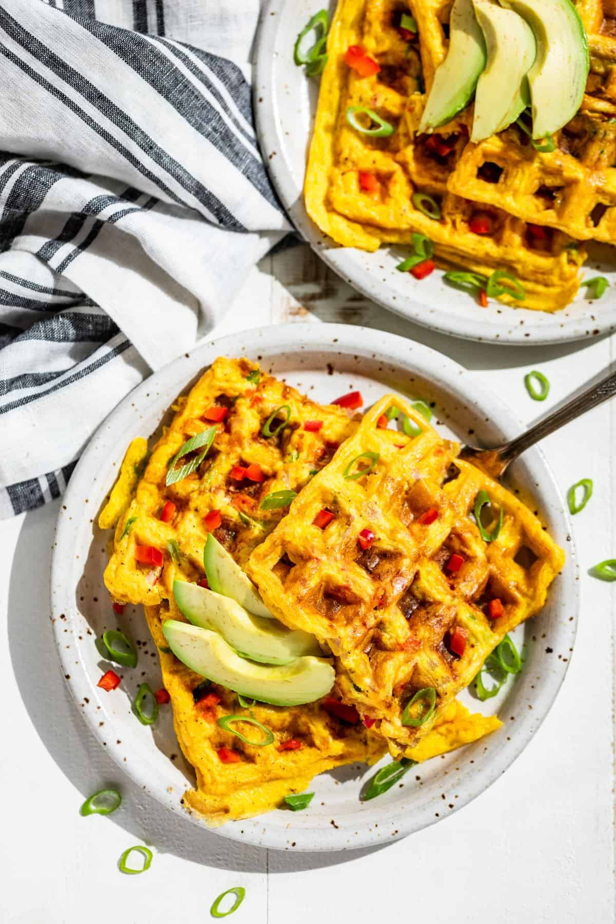 Straight down view of two plates with Western Omelet Waffles topped with sliced green onions and avocado.