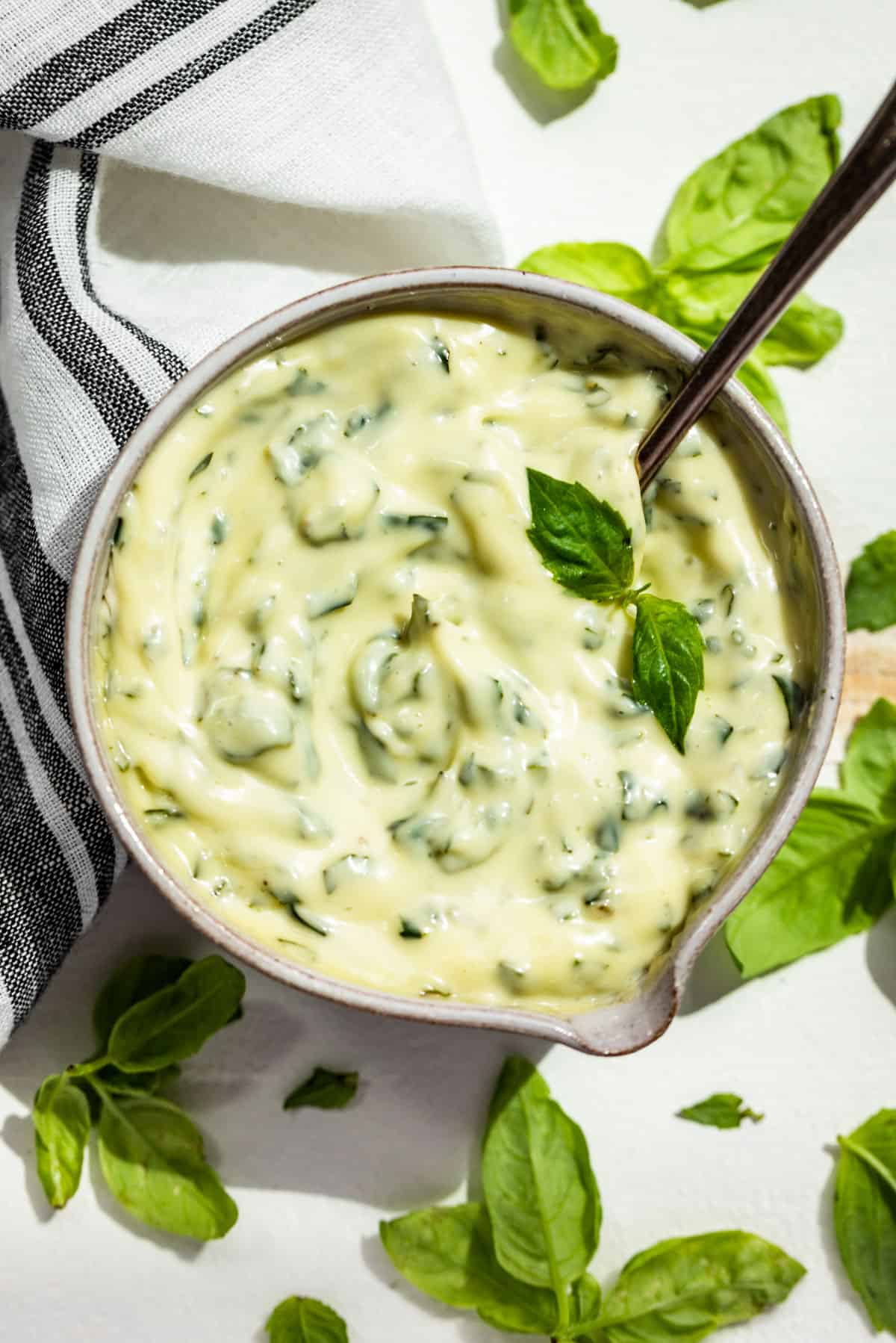 Straight down view of Basil Aioli in a pottery bowl with a silver spoon and basil leaves around it.