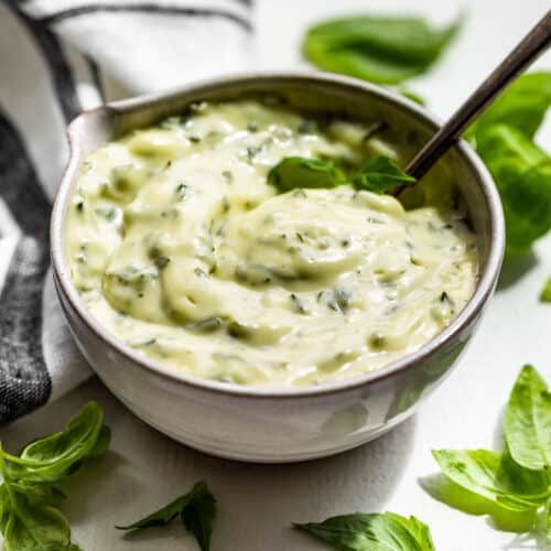 Side view of Basil Aioli in a pottery bowl with a silver spoon and basil leaves around it.