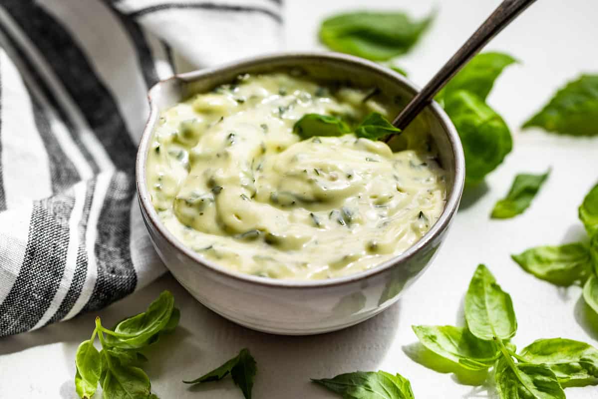 Side view of Basil Aioli in a pottery bowl with a silver spoon and basil leaves around it.