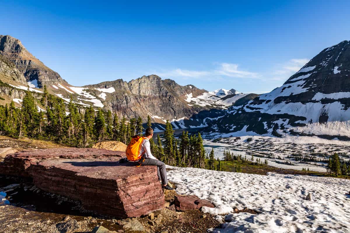 Man with an orange backpack sitting on a large rock overlooking Hidden Lake in Glacier National Park.