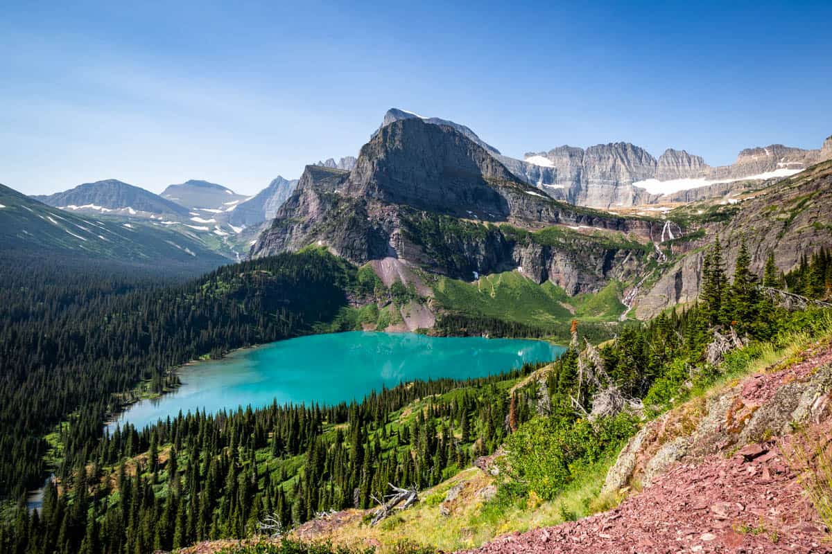 Aqua blue lake called Grinnell Lake with a mountain peak above it and red rock in the foreground.