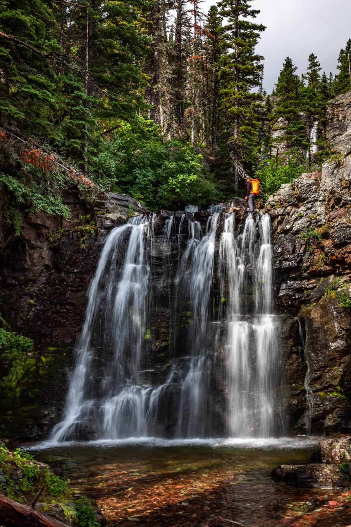 Large waterfall over dark brown rocks with trees around it and a man in an orange backpack standing at the top of the falls.
