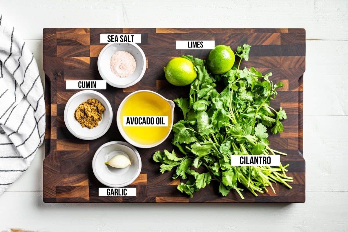 Cilantro, lime, avocado oil, garlic, cumin, and sea salt on a wood cutting board.