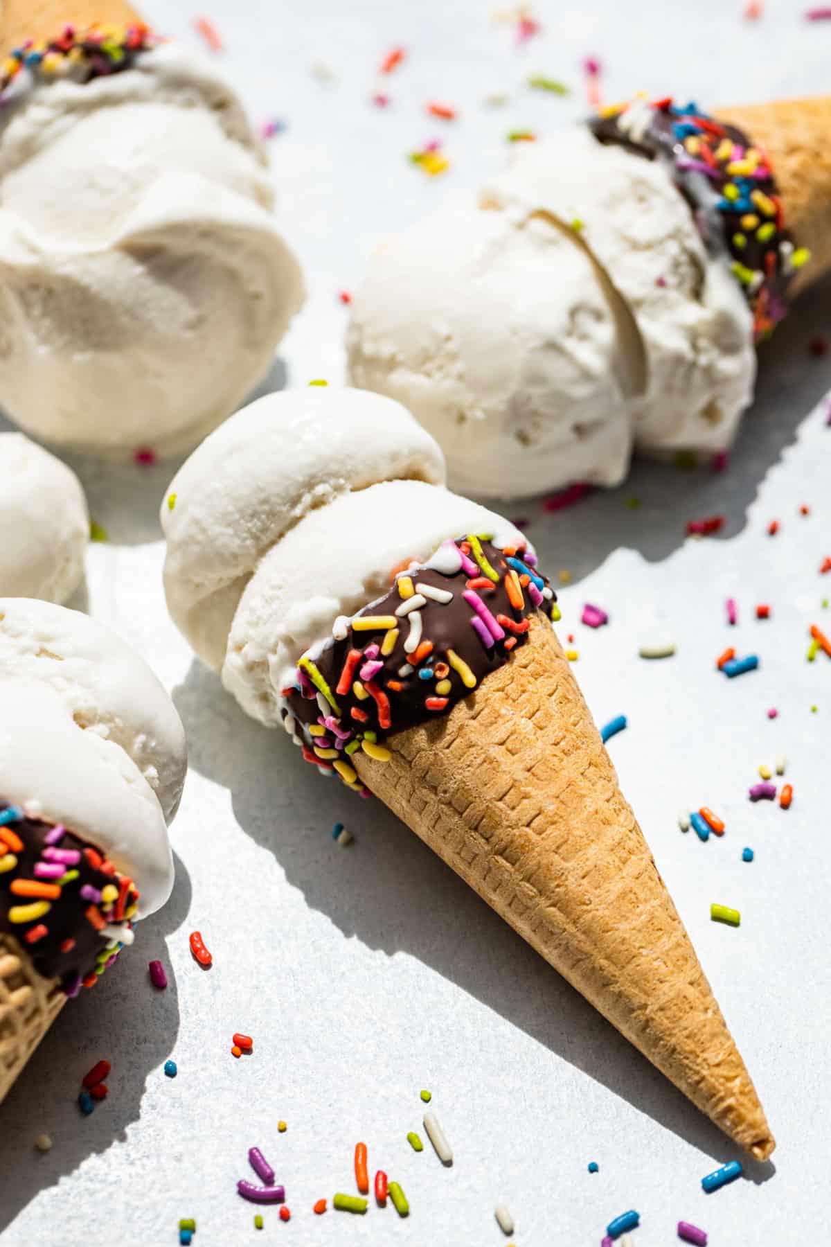 Coconut Ice Cream in chocolate dipped cones with sprinkles around them on a blue background.