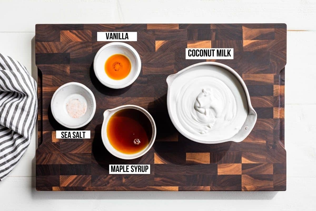 Coconut milk, maple syrup, vanilla extract, and sea salt in small bowls on a wood cutting board.