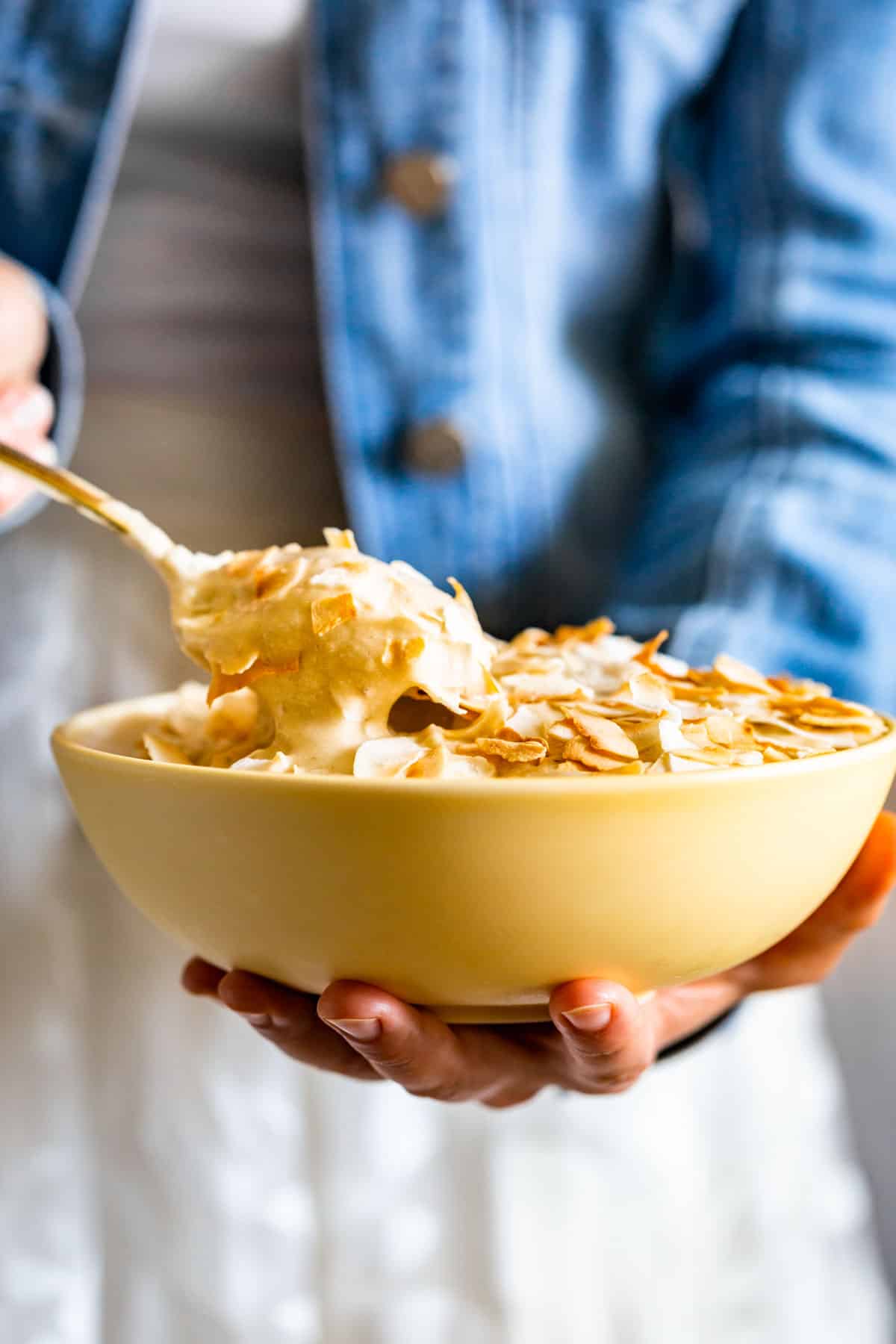 A yellow bowl with Coconut Banana Nice Cream topped with toasted coconut with a person in a blue jacket in the background.