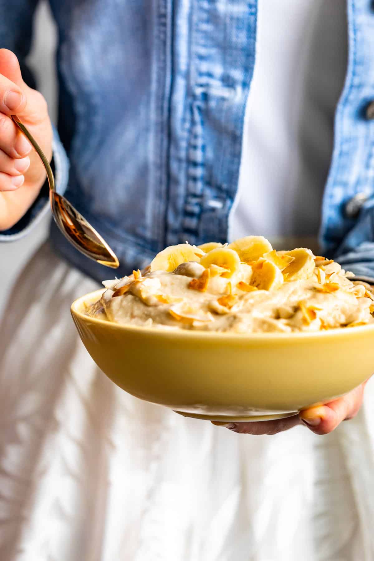 A yellow bowl filled with Creamy Coconut Banana Nice Cream with a person in a blue jacket holding the bowl.