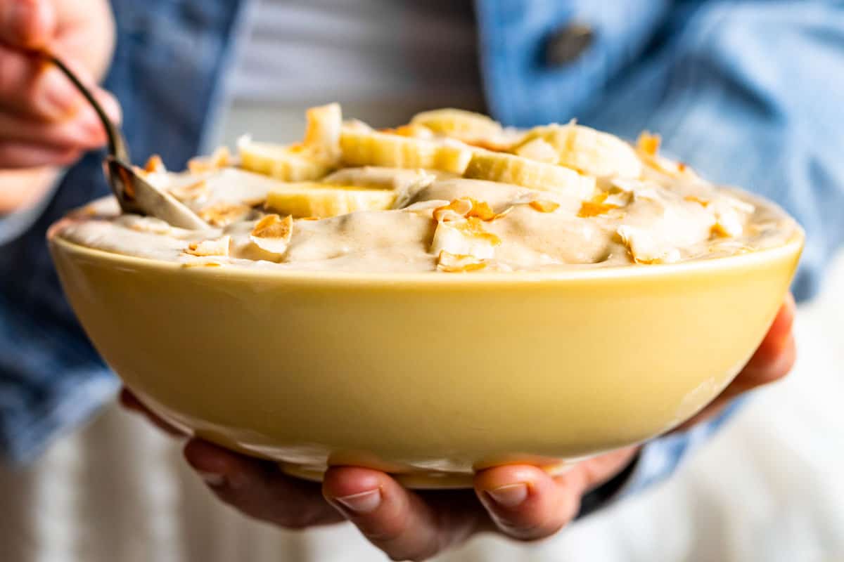 A yellow bowl filled with Coconut Banana Nice Cream with a person in a blue jacket scooping some out.