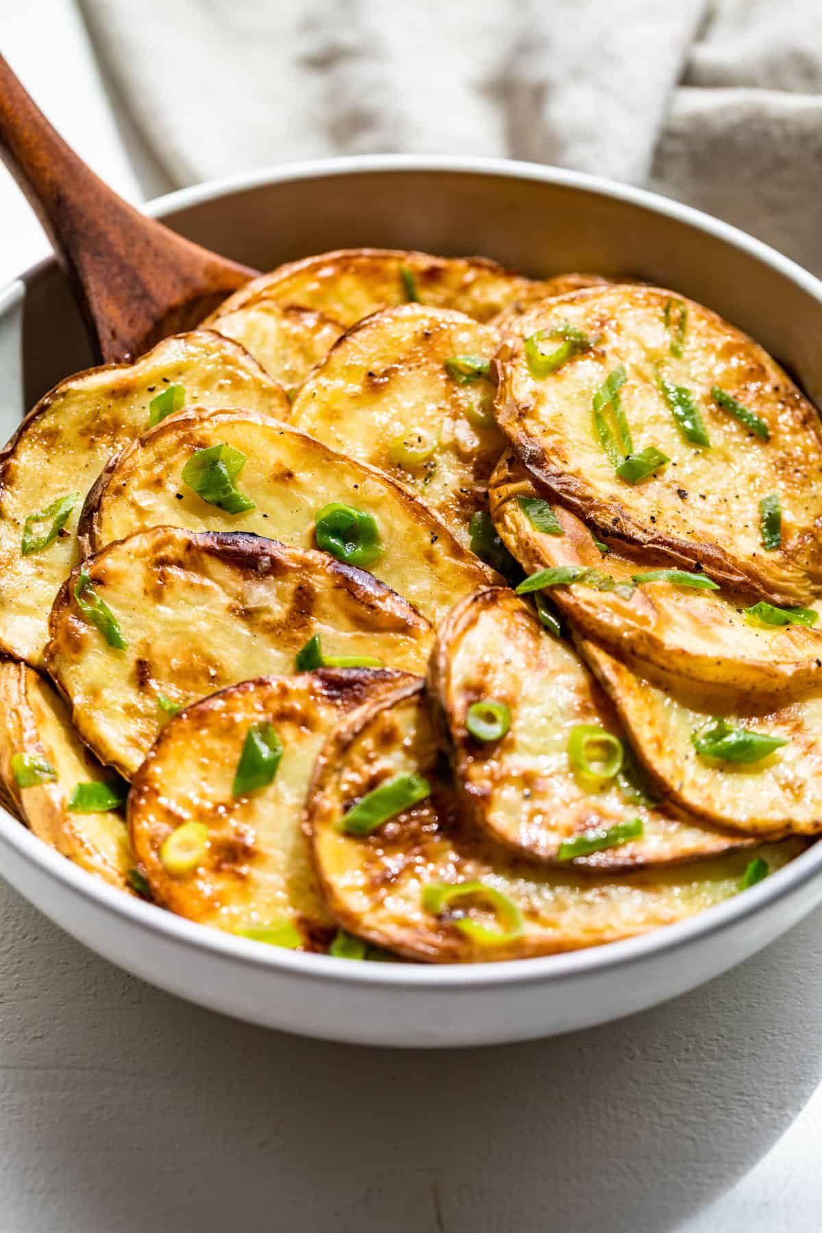 A large white bowl with grilled sliced potatoes topped with green onions with a wood spoon.