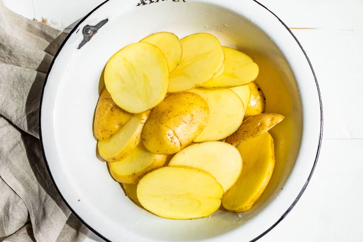 A large white enamel bowl filled with sliced potatoes coated in avocado oil.