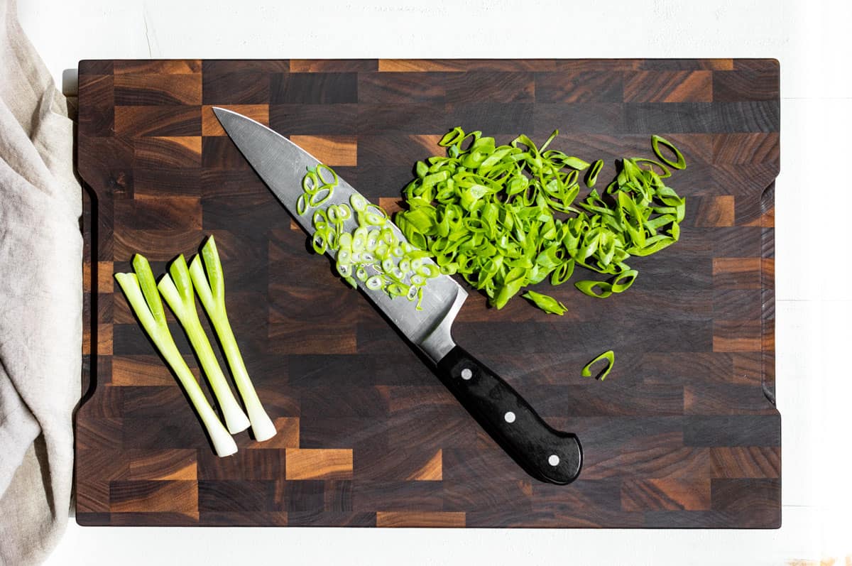 Slicing green onions with a large chef's knife on a wood cutting board.