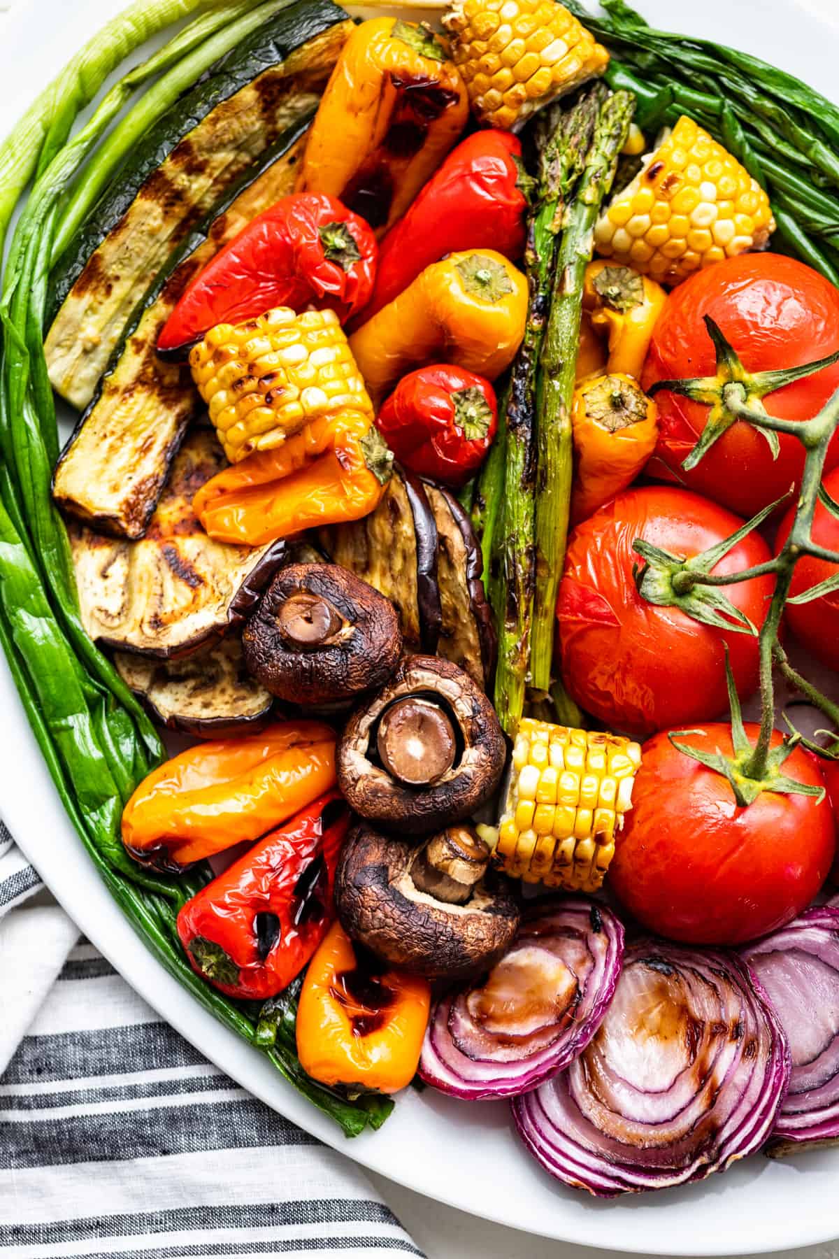 Lots of grilled veggies on a white platter, corn on the cob, tomatoes, mushrooms, sliced red onions, gypsy sweet peppers, zucchini, and asparagus.