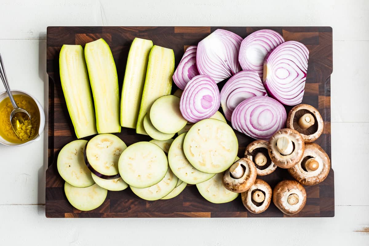 Mushrooms, sliced eggplant, zucchini, and red onions on a wood cutting board.
