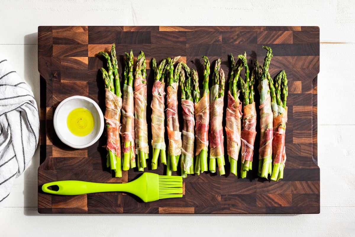 Prosciutto Wrapped Asparagus on a wooden cutting board being brushed with olive oil with a green silicone brush.