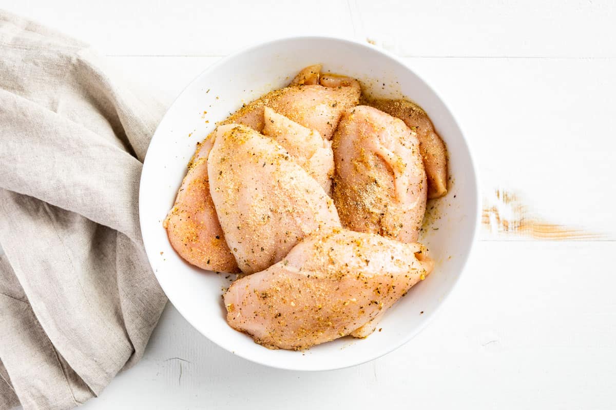 Raw chicken breasts in a white bowl being tossed together with olive oil, seasonings, and sea salt.