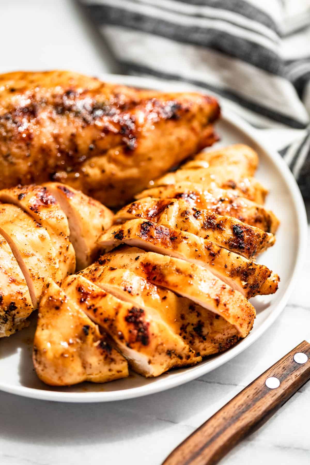 Side view of sliced Smoked Chicken Breasts on a white plate with a wood handled knife on the side and blue and white striped linen in the background.