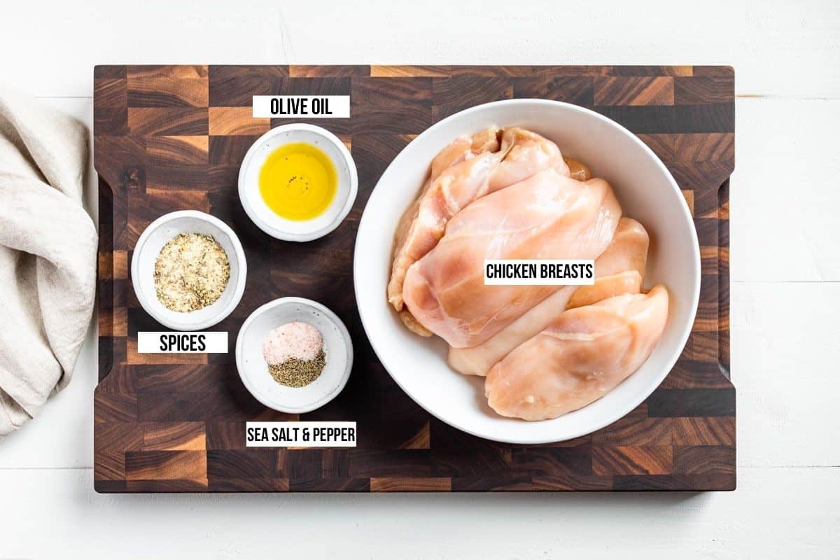 Chicken breasts in a white bowl, with olive oil , seasoning, and sea salt in bowls on the side all on a wood cutting board.