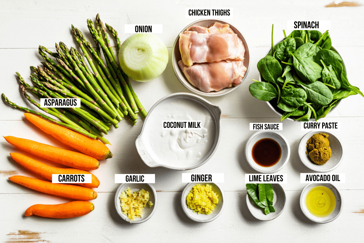 Asparagus, carrots, onion, ginger, garlic, curry pasta, chicken thighs, spinach, lime leaves, fish sauce, and coconut milk on a wooden cutting board.