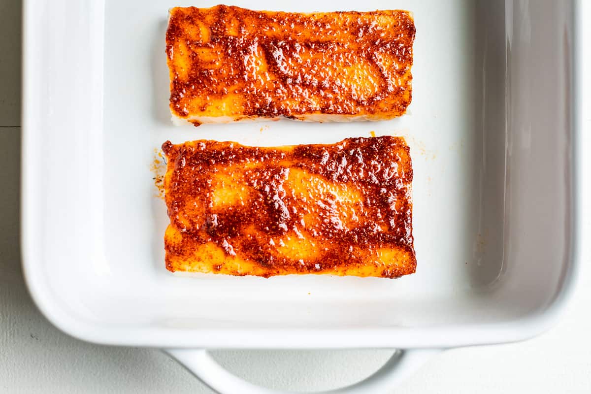 Fish fillets with the spice mixture smeared on top in a white baking dish.