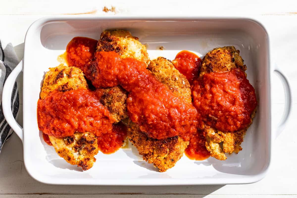 Pan fried chicken cutlets in a white baking dish being topped with tomato sauce.