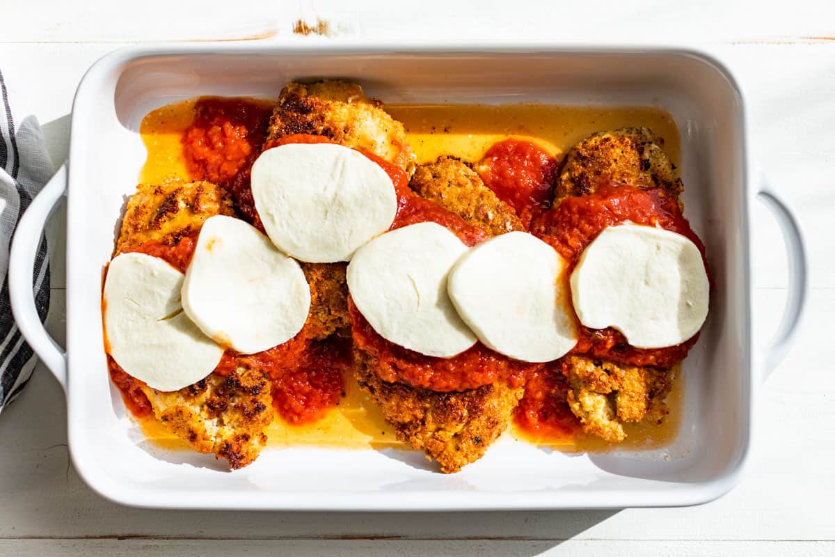 Adding the fresh mozzarella slice to the chicken cutlets in a white baking dish.