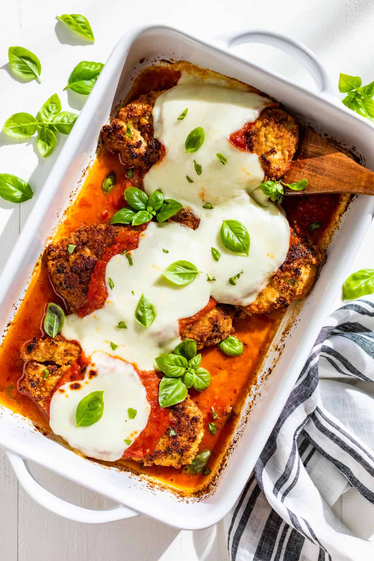 Four pieces of Almond Flour Chicken Parmesan topped with sprigs of basil in a white baking dish.