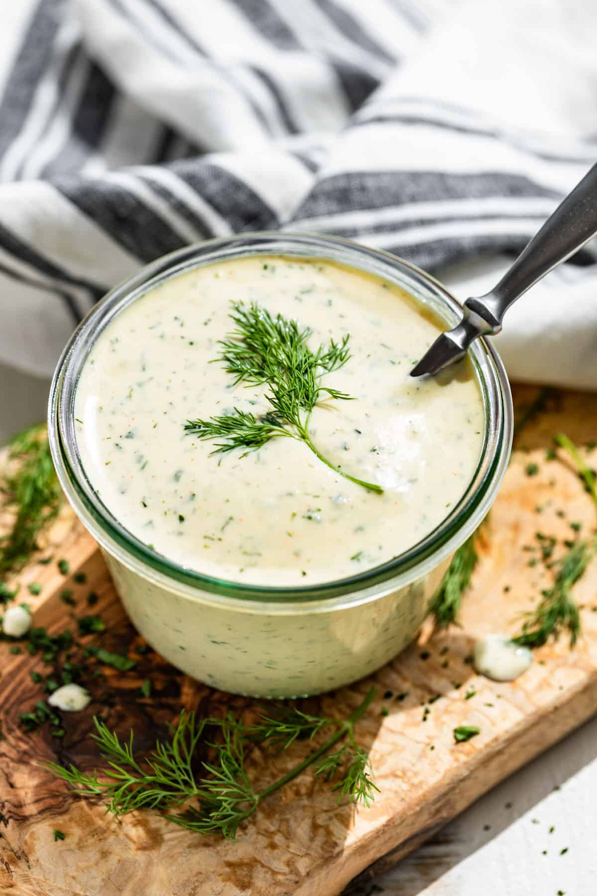 Ranch dressing in a glass jar with a silver spoon in it on a wooden board with chopped dill around it.