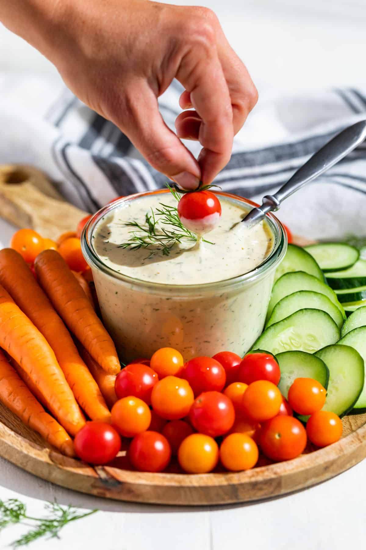 A hand dipping a cherry tomato in a container of ranch dressing surrounded by cut veggies.