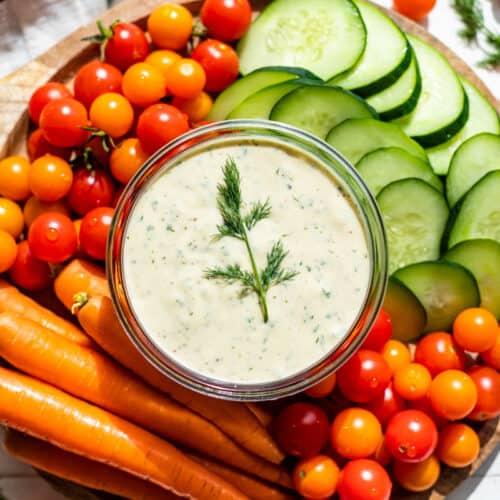 Dairy Free Ranch Dressing in a glass container on a wooden board surrounded by carrots, cherry tomatoes, and sliced cucumbers.