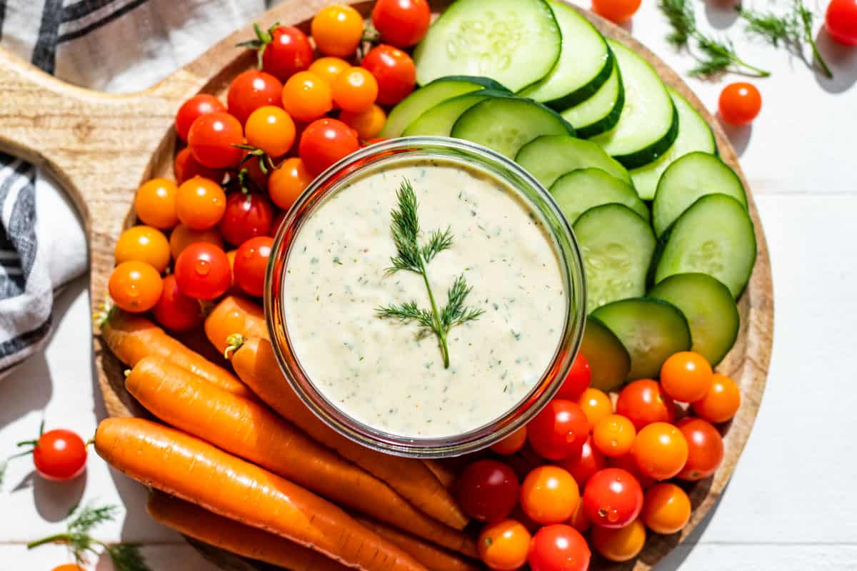 Dairy Free Ranch Dressing in a glass container on a wooden board surrounded by carrots, cherry tomatoes, and sliced cucumbers.