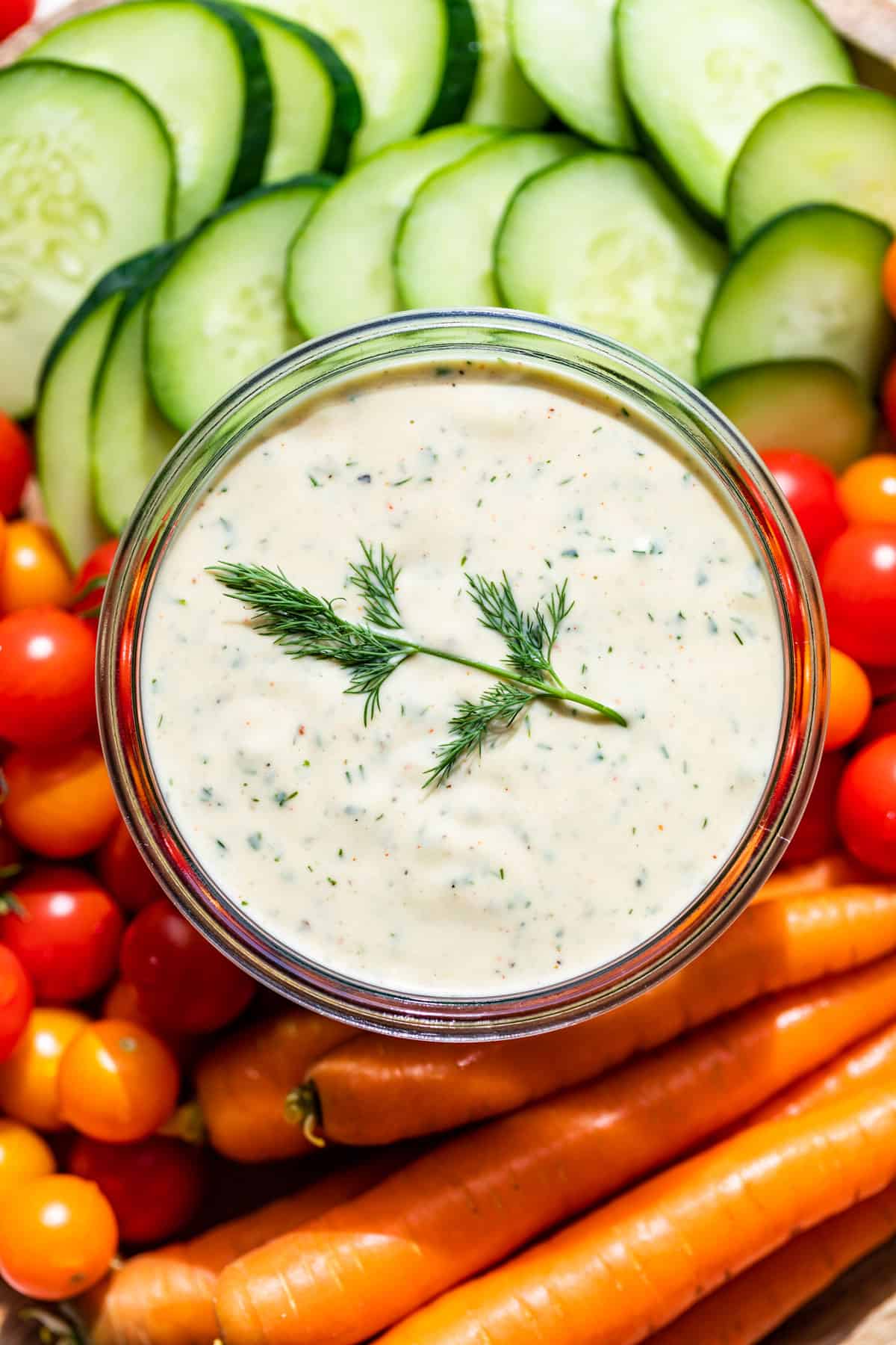 Close up view of Dairy Free Ranch Dressing in a glass jar with a sprig of dill on top surrounded by cucumber slices, carrots, and cherry tomatoes.
