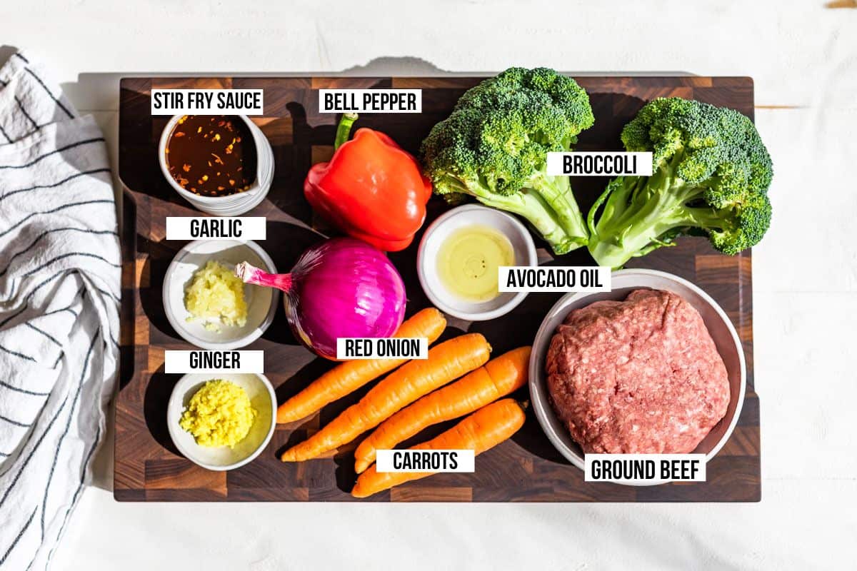 All the ingredients for the Ground Beef Stir Fry on a wood cutting board. Broccoli, bell pepper, carrots, onion, ground beef, garlic, ginger, and stir fry sauce.