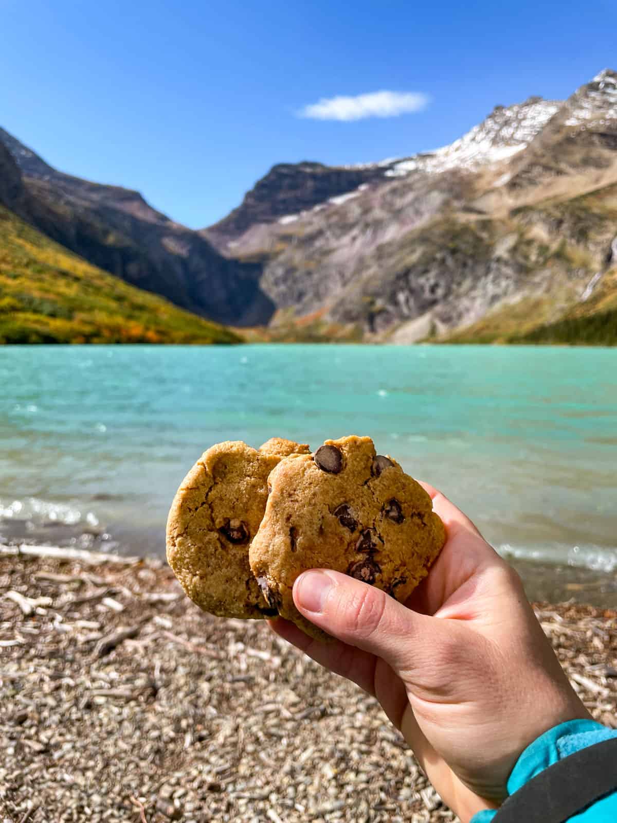 Two chocolate chip cookies held in a hand with the turquoise lake and mountains in the background.