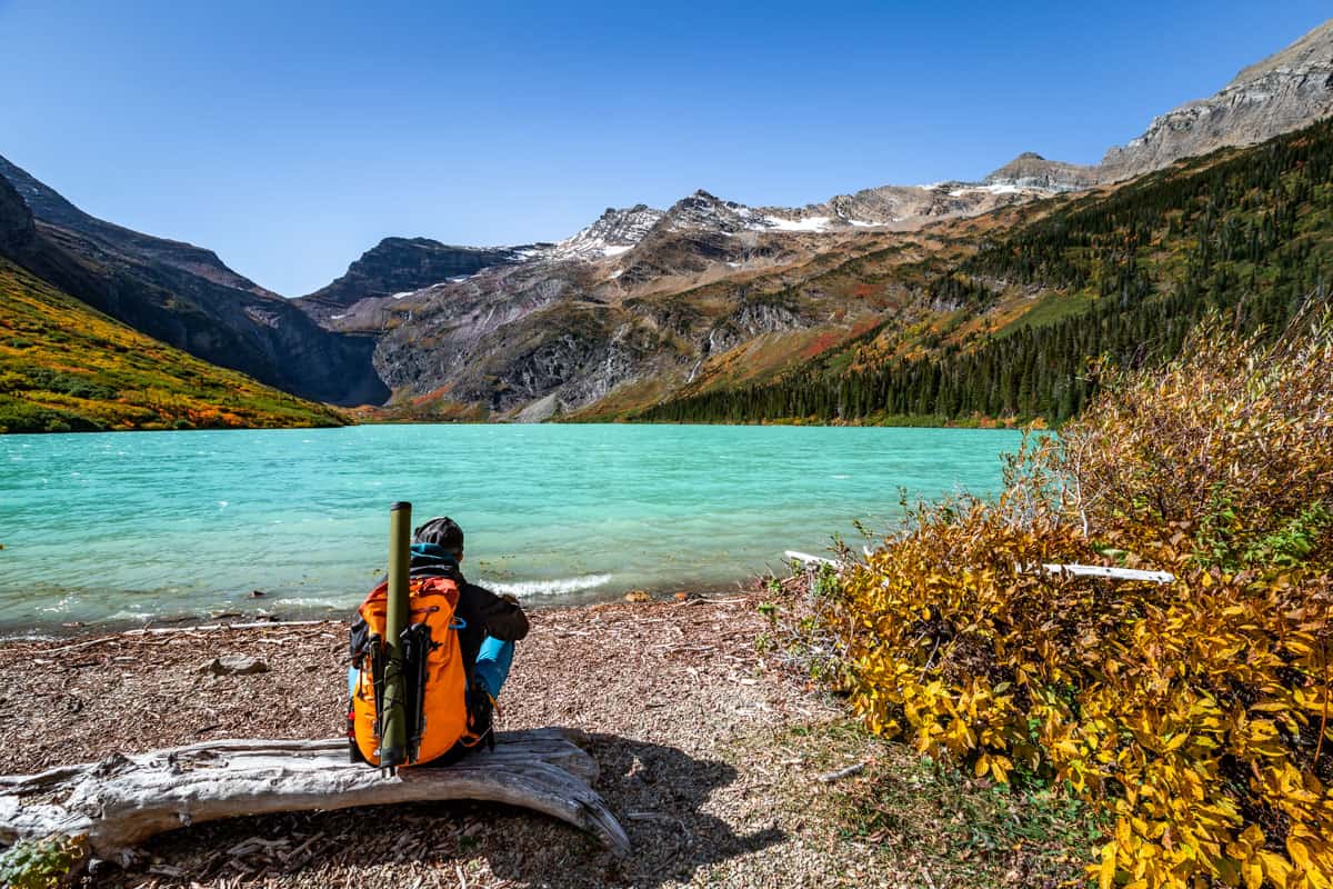 A man with an orange backpack sitting on a log looking at a turquoise lake with mountains in the background.