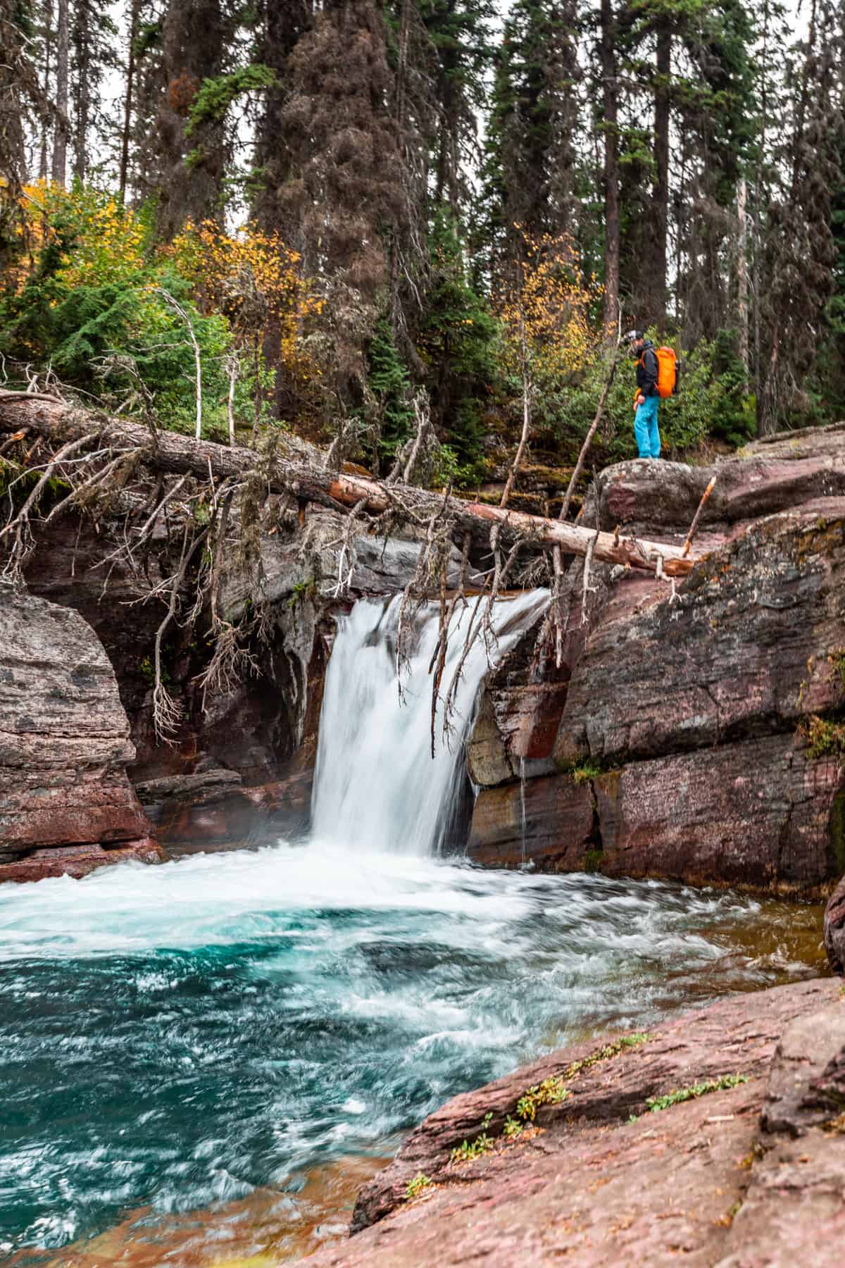 A turquoise colored waterfall with red rock on either side and a man with an orange back pack standing at the top.