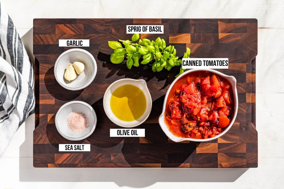 All the ingredients for Marinara Sauce on a wood cutting board, garlic, tomatoes, olive oil, sea salt, and a sprig of basil.