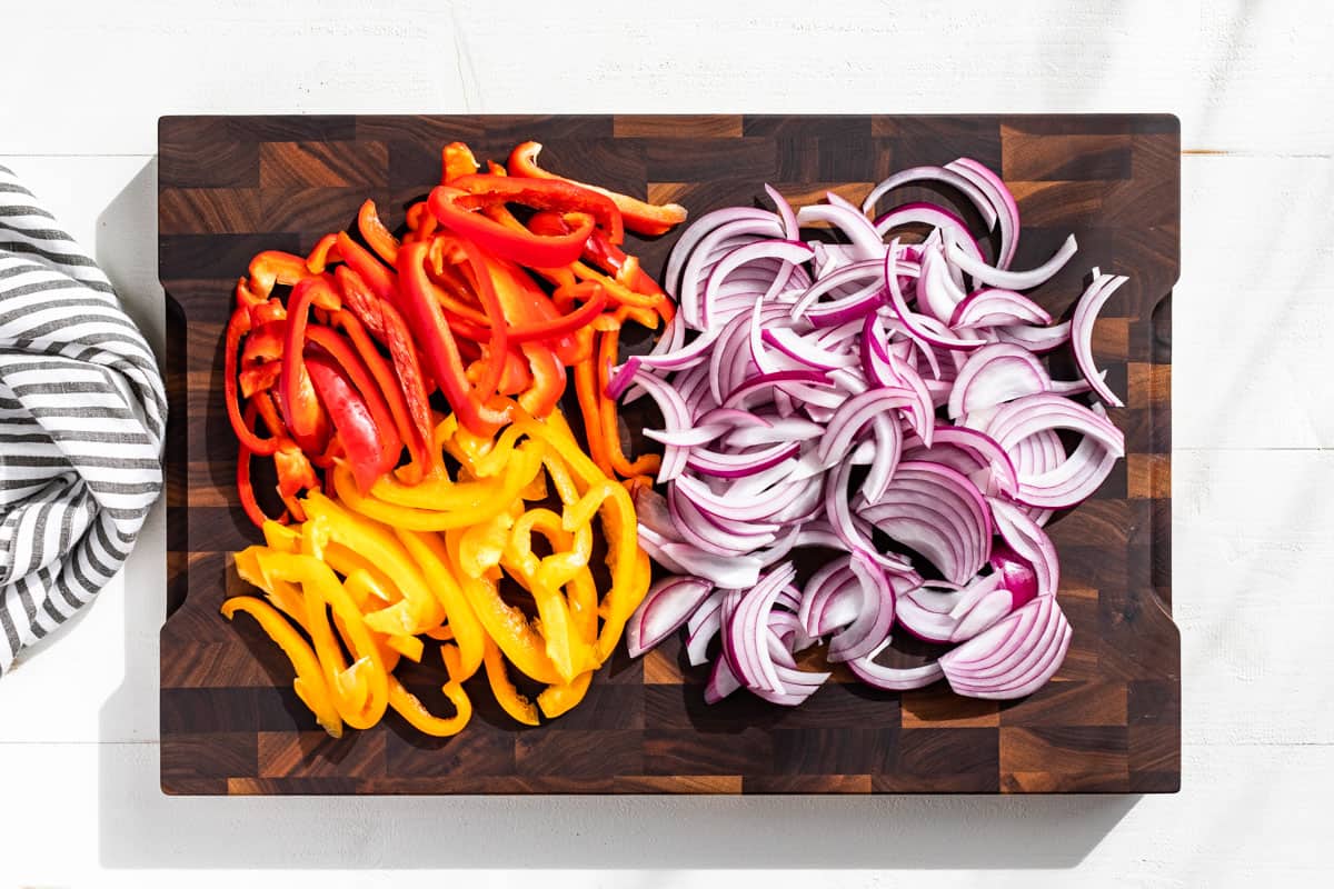Sliced red and yellow bell peppers and sliced red onions on a wood cutting board.