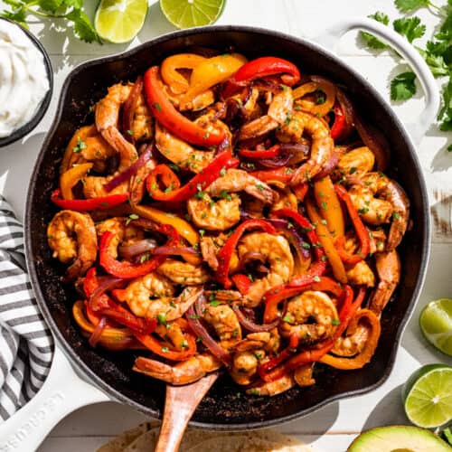 Straight down view of finished Shrimp Fajitas in a white skillet with avocado, salsa, and sour cream on the side.