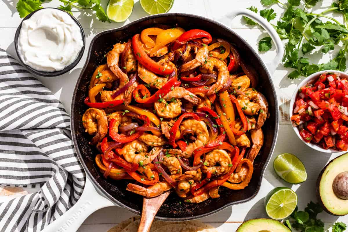 Straight down view of finished Shrimp Fajitas in a white skillet with avocado, salsa, and sour cream on the side.