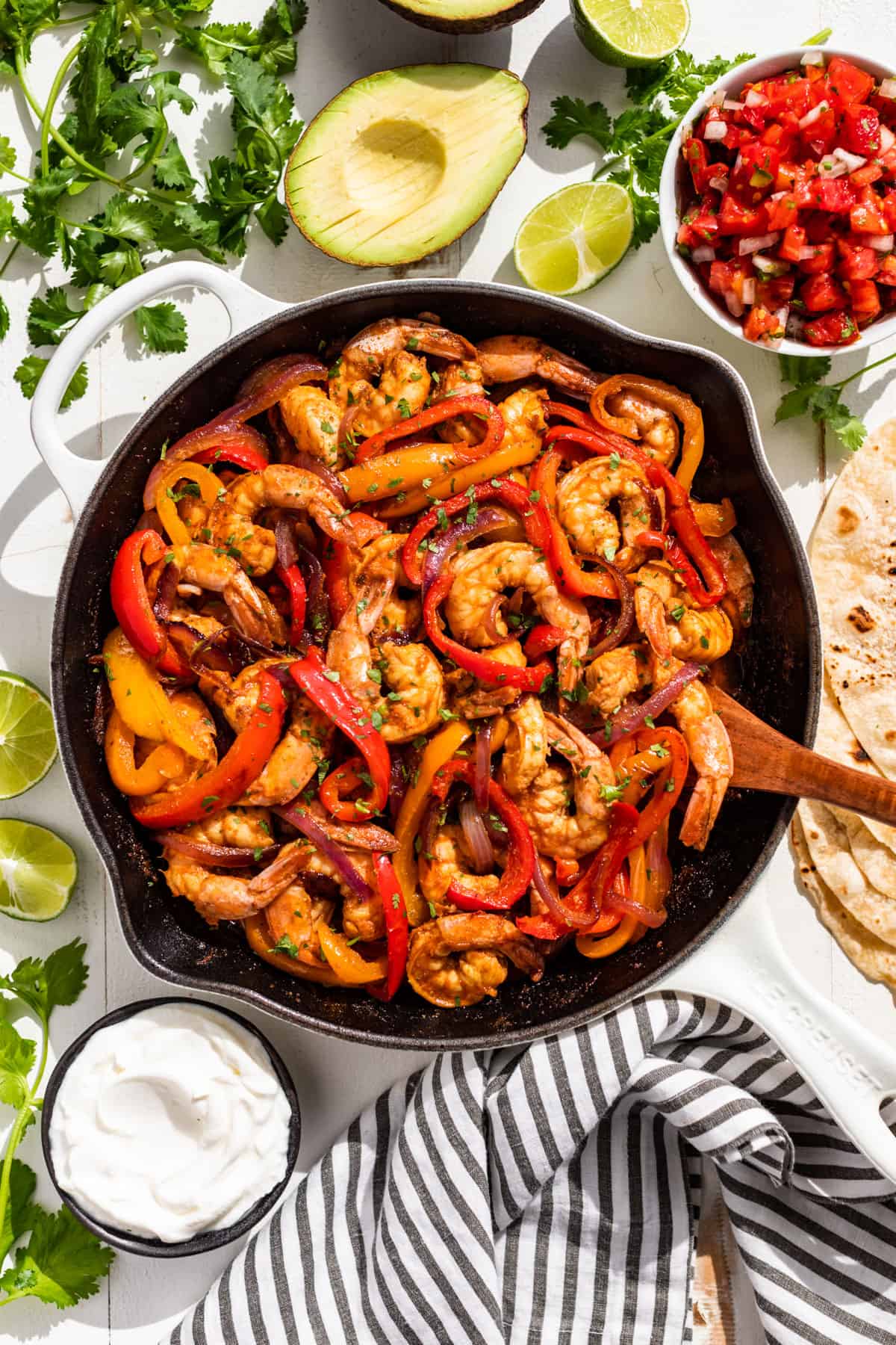 Shrimp Fajitas in a white skillet with sour cream, salsa, avocados, and cilantro on the side.