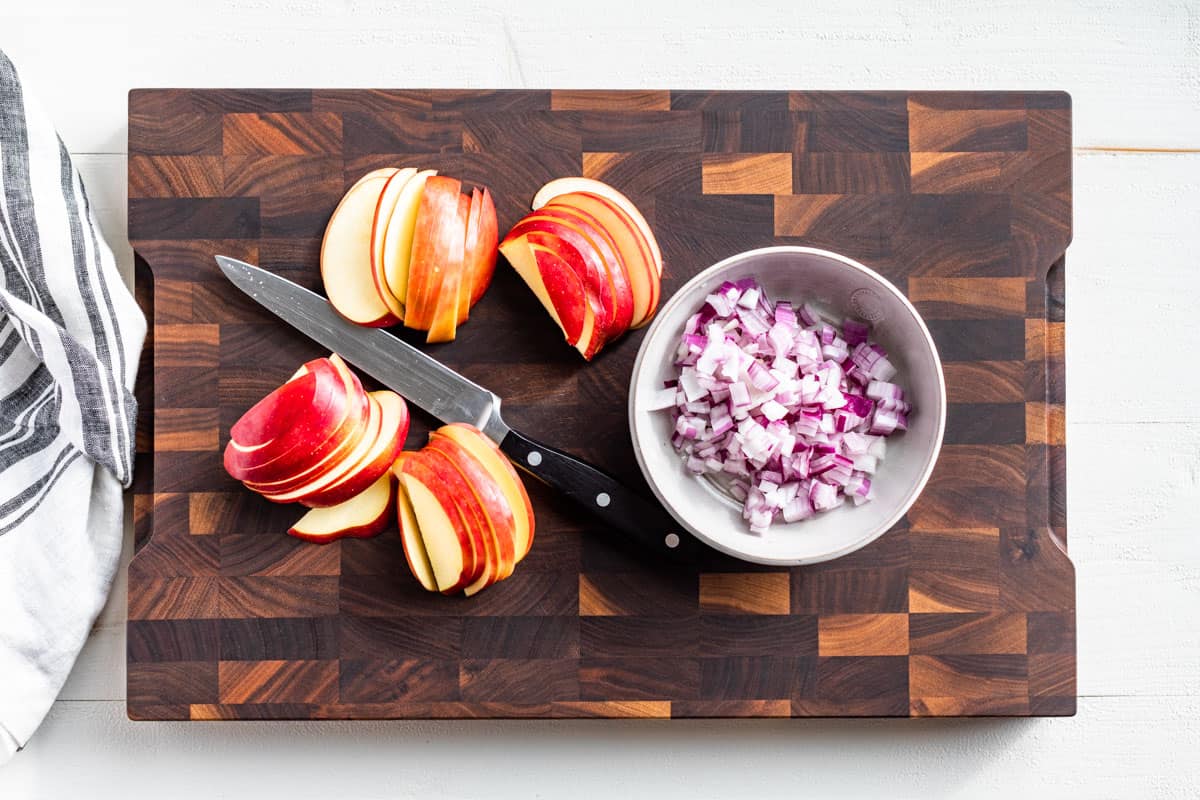 Diced red onion in a bowl and sliced apples on a wood cutting board.