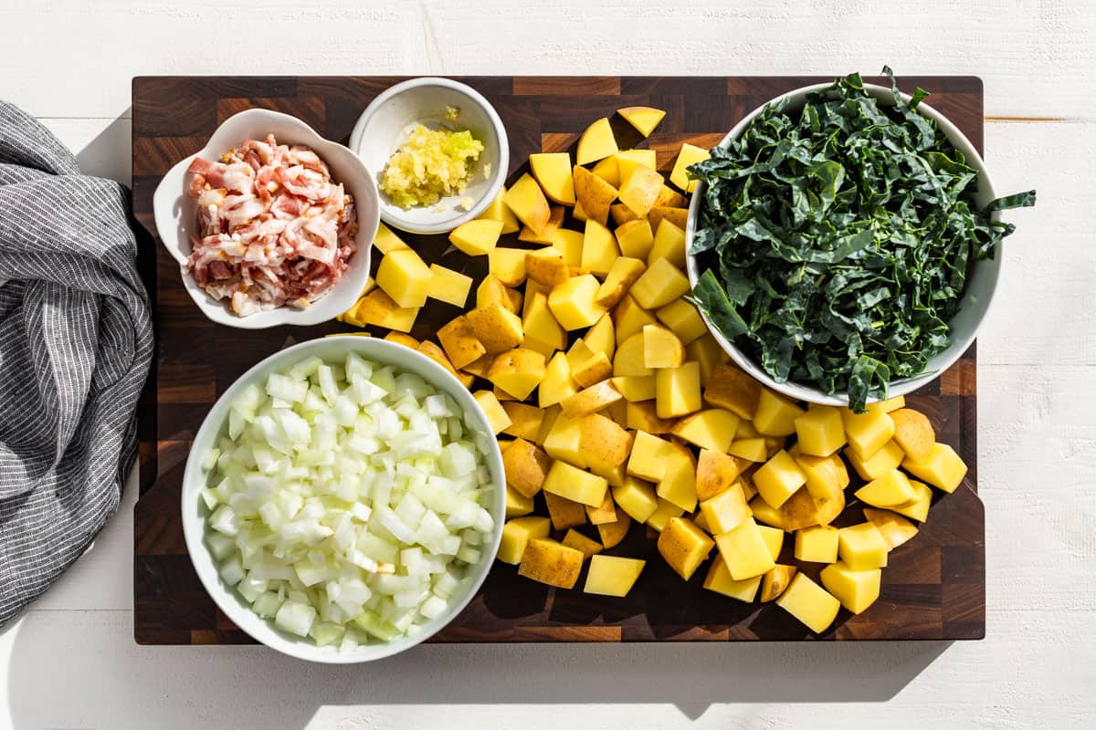 Cut up bacon, diced onion, minced garlic, diced potatoes, and sliced kale on a wood cutting board.