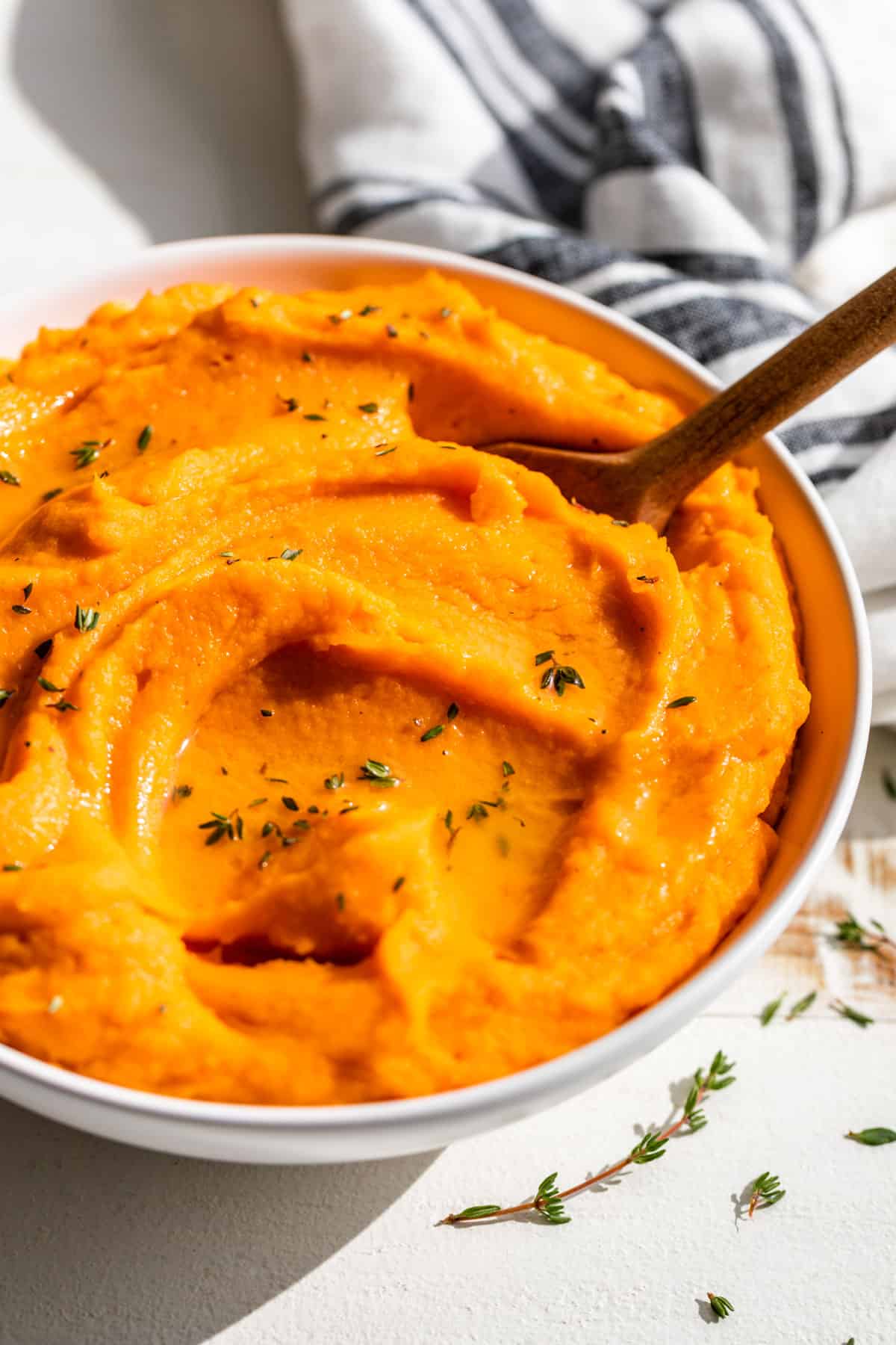 Side view of Mashed Sweet Potatoes in a white bowl with a wood spoon and blue and white striped linen in the background.