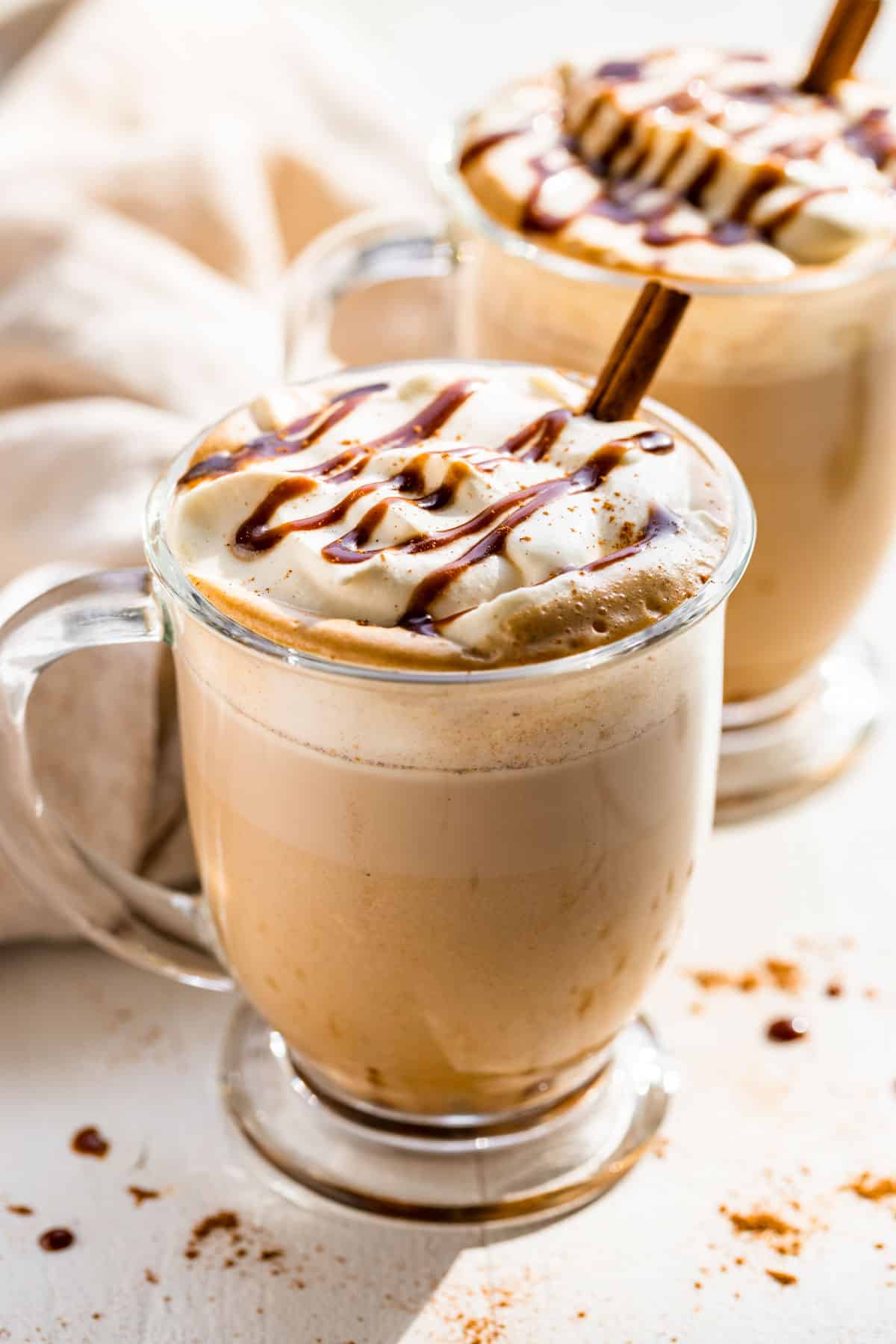 Pumpkin Chai Latte in two glass mugs on a white background and topped with whipped cream, caramel, and a cinnamon stick.