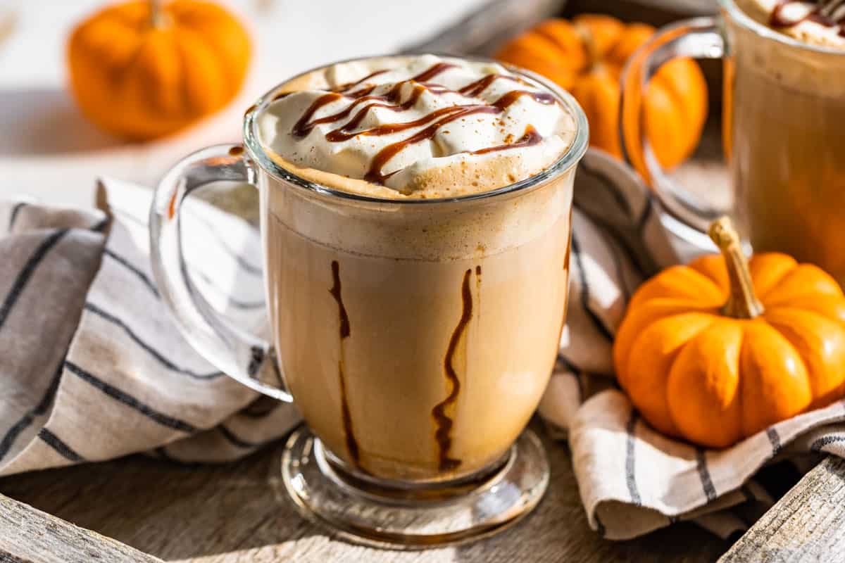 Two mugs of Pumpkin Chai Latte topped with whipped cream and caramel sauce with mini pumpkins around the glass mugs.