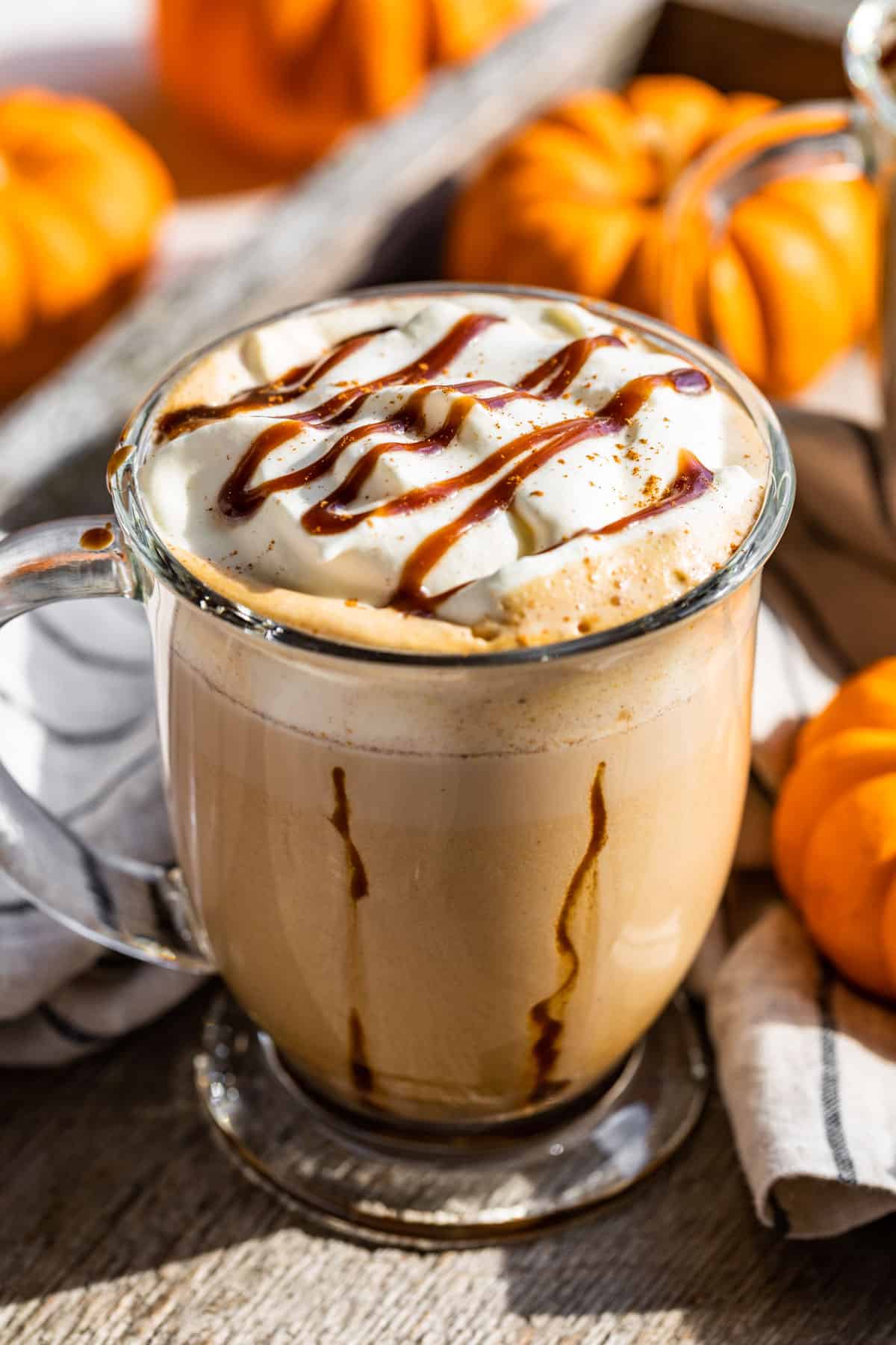 Pumpkin Chai Latte in a glass mug with caramel stripes down the sides of the mug and topped with whipped cream and caramel with mini pumpkins around it.