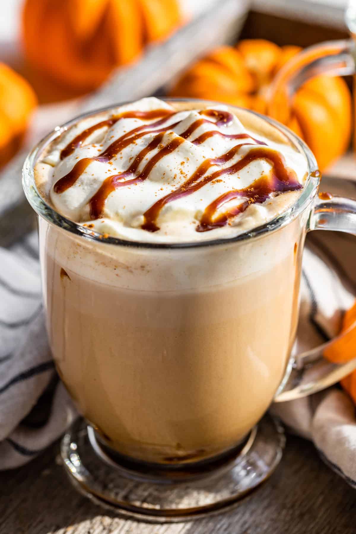 A glass mug filled with Pumpkin Chai Latte and topped with whipped cream and caramel sauce with pumpkins in the background.