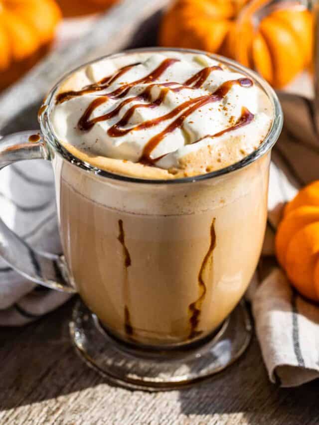 Pumpkin Chai Latte in a glass mug with caramel stripes down the sides of the mug and topped with whipped cream and caramel with mini pumpkins around it.