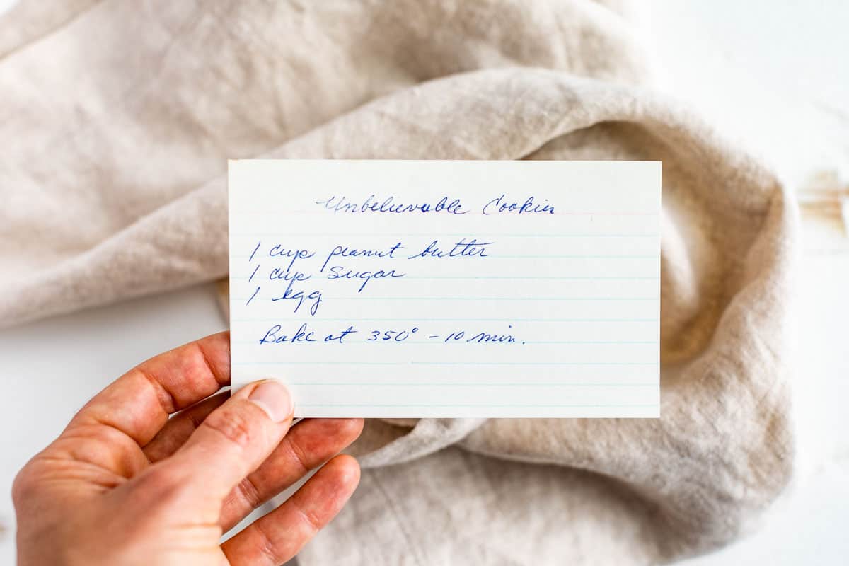 A hand holding a very old hand written recipe for 3 Ingredient Peanut Butter Cookies titled 'Unbelievable Cookies'.